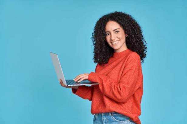 a woman with curly hair standing in front of a blue backdrop holding a laptop with her right hand and typing with her left hand