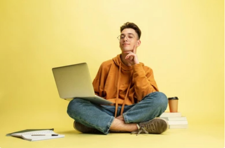 Young man in a casual orange hoodie and blue jeans sitting cross-legged on the floor with a laptop on his lap, looking thoughtful against a yellow background, surrounded by books and a coffee cup.