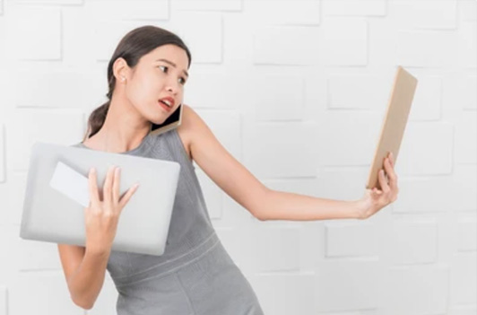 A woman in a gray dress looks stressed while juggling a laptop and a book, against a white brick wall background.