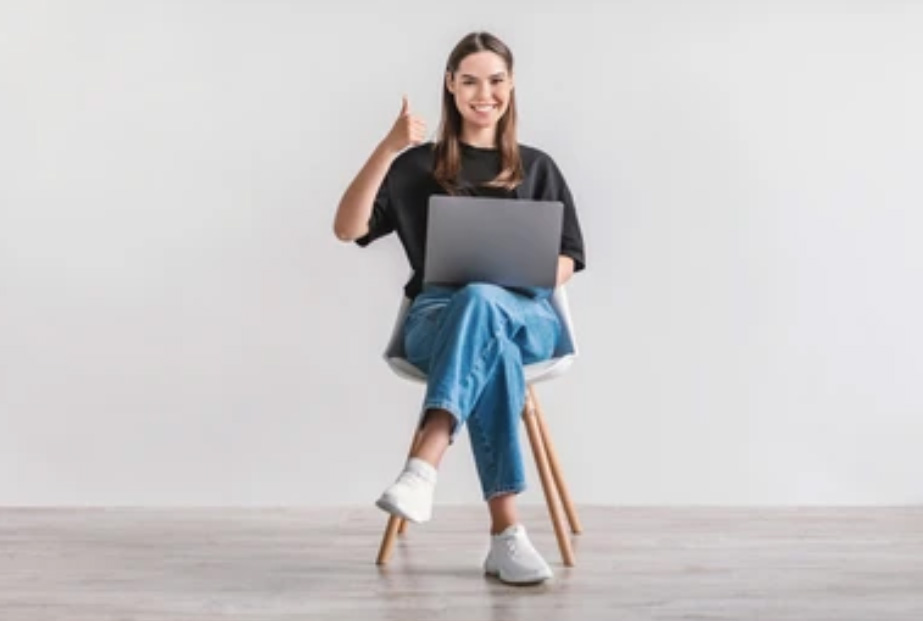 A female sitting on a chair with her thumb up and a laptop on her lap